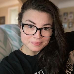 An image of Professor Arianna Stone smiling at the camera. She is wearing black glasses, a black shirt with some white writing, and her hair is down and pulled over 1 shoulder.